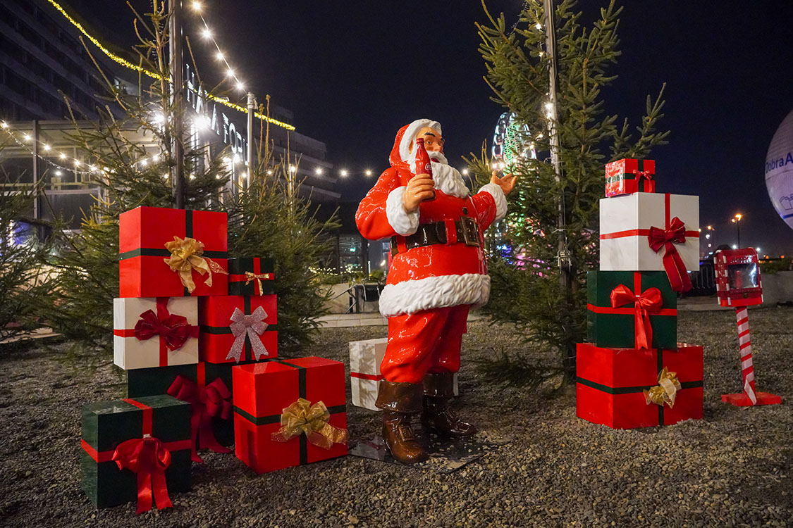 Jeder kann der Weihnachtsmann sein: Coca-Colas Weihnachtskampagne mit Weihnachtsmannfiguren 2 Figurka Mikołaja trzymającego butelkę napoju w otoczeniu świątecznie udekorowanych prezentów i małych drzewek iglastych oświetlonych lampkami nocą w plenerze.