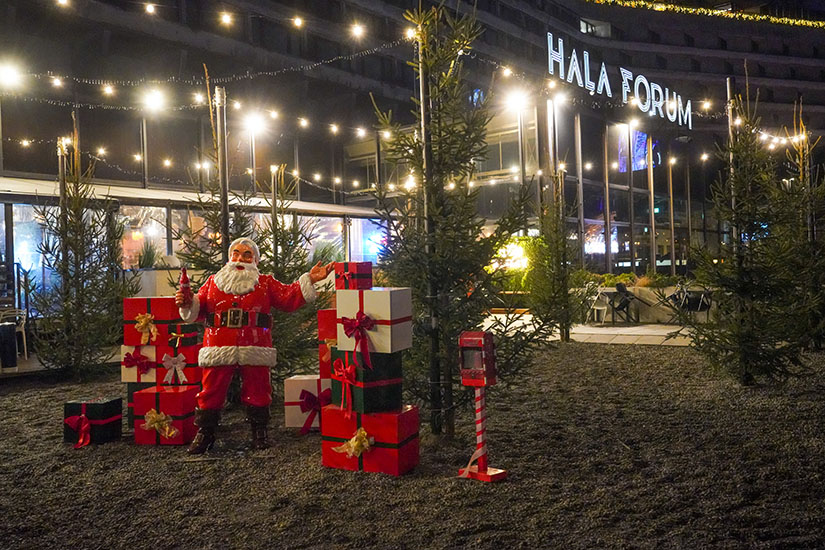 Jeder kann der Weihnachtsmann sein: Coca-Colas Weihnachtskampagne mit Weihnachtsmannfiguren 4 Rankiem na zewnątrz hali Forum ozdobionej światełkami, stoi duża figurka Świętego Mikołaja otoczona wysokimi choinkami i kolorowymi, ozdobnymi prezentami; w tle szklana fasada i neonowa nazwa Hala Forum.