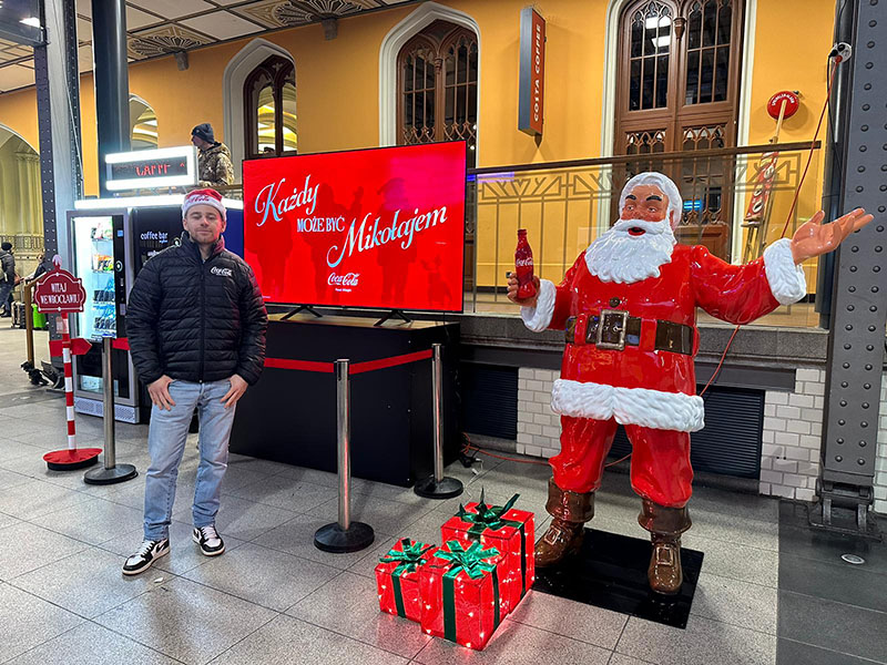 Jeder kann der Weihnachtsmann sein: Coca-Colas Weihnachtskampagne mit Weihnachtsmannfiguren 5 Mężczyzna w czapce Mikołaja stoi obok dużej figurki Świętego Mikołaja trzymającego butelkę Coca-Coli, obok znajdują się podświetlane prezenty, a za nimi czerwony ekran z napisem