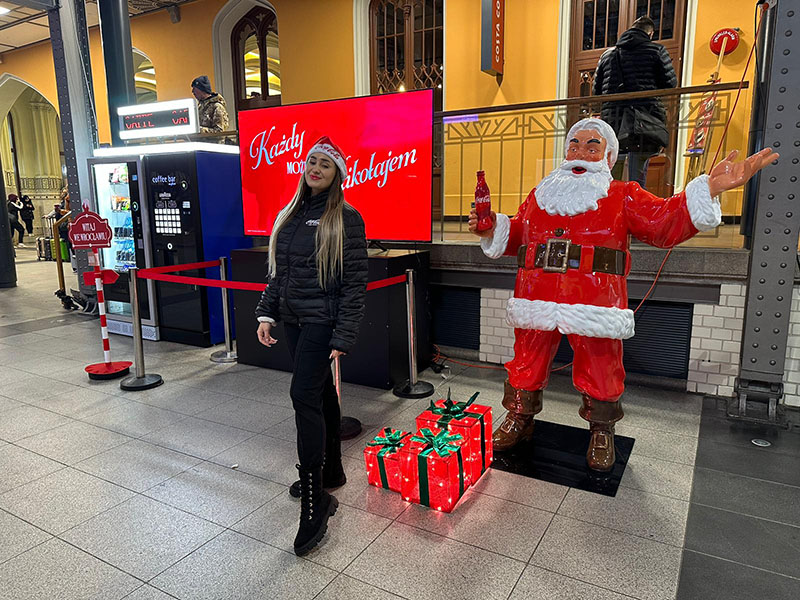 Jeder kann der Weihnachtsmann sein: Coca-Colas Weihnachtskampagne mit Weihnachtsmannfiguren 6 Kobieta w czarnej kurtce i czapce świętego Mikołaja stoi na dworcu obok dużej figurki Mikołaja trzymającej butelkę Coca-Coli i podświetlonych prezentów w czerwonym papierze z zielonymi kokardami.