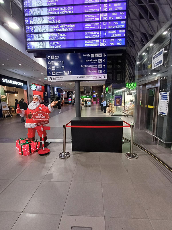Jeder kann der Weihnachtsmann sein: Coca-Colas Weihnachtskampagne mit Weihnachtsmannfiguren 7 Wnętrze dworca z tablicą odjazdów pociągów na górze, na dole figura Mikołaja z prezentami i napisem Witaj w Poznaniu oraz sklepami Starbucks i Żabka w tle.