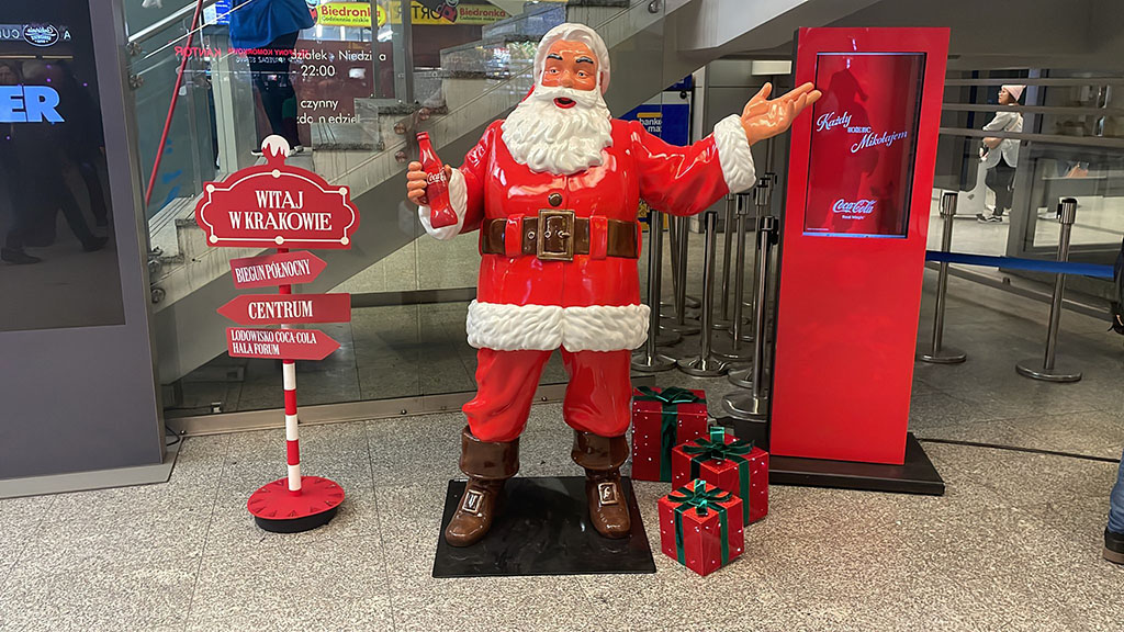 Jeder kann der Weihnachtsmann sein: Coca-Colas Weihnachtskampagne mit Weihnachtsmannfiguren 8 Figurka Mikołaja w czerwonym stroju trzymającego butelkę Coca-Coli, stojąca obok czerwonego stojaka z ekranem oraz tabliczki z napisem Witaj w Krakowie i kierunkami do Bieguna Północnego, centrum i lodowiska Coca-Coli, a także przedmiotów wyglądających jak prezenty na podłodze, w nowoczesnym pomieszczeniu z szarym posadzką i przeszklonym wejściem w tle