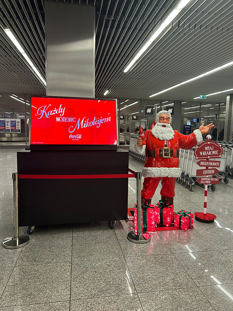 Jeder kann der Weihnachtsmann sein: Coca-Colas Weihnachtskampagne mit Weihnachtsmannfiguren 9 Figurka Świętego Mikołaja z butelką Coca-Coli i dekoracyjnymi prezentami przy informacji lotniskowej, obok stoi czerwony ekran z napisem