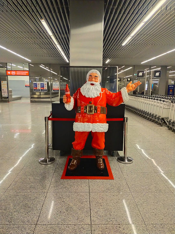 Jeder kann der Weihnachtsmann sein: Coca-Colas Weihnachtskampagne mit Weihnachtsmannfiguren 10 Figurka Mikołaja w czerwonym stroju z białym futrem stojąca na czarno-czerwonym podłożu, trzymająca czerwoną butelkę, w nowoczesnym hali z metalowymi sufitami i rzędem wózków po prawej stronie