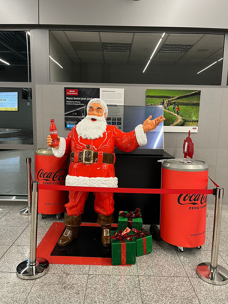 Jeder kann der Weihnachtsmann sein: Coca-Colas Weihnachtskampagne mit Weihnachtsmannfiguren 11 Figurka Mikołaja w czerwonym stroju trzyma butelkę Coca-Coli, stoi między dwoma dużymi pojemnikami przypominającymi puszki Coca-Coli, obok leżą dwa zielone prezenty z czerwonymi wstążkami, całość znajduje się w pomieszczeniu z szarymi ścianami i dużym lustrem za figurką.