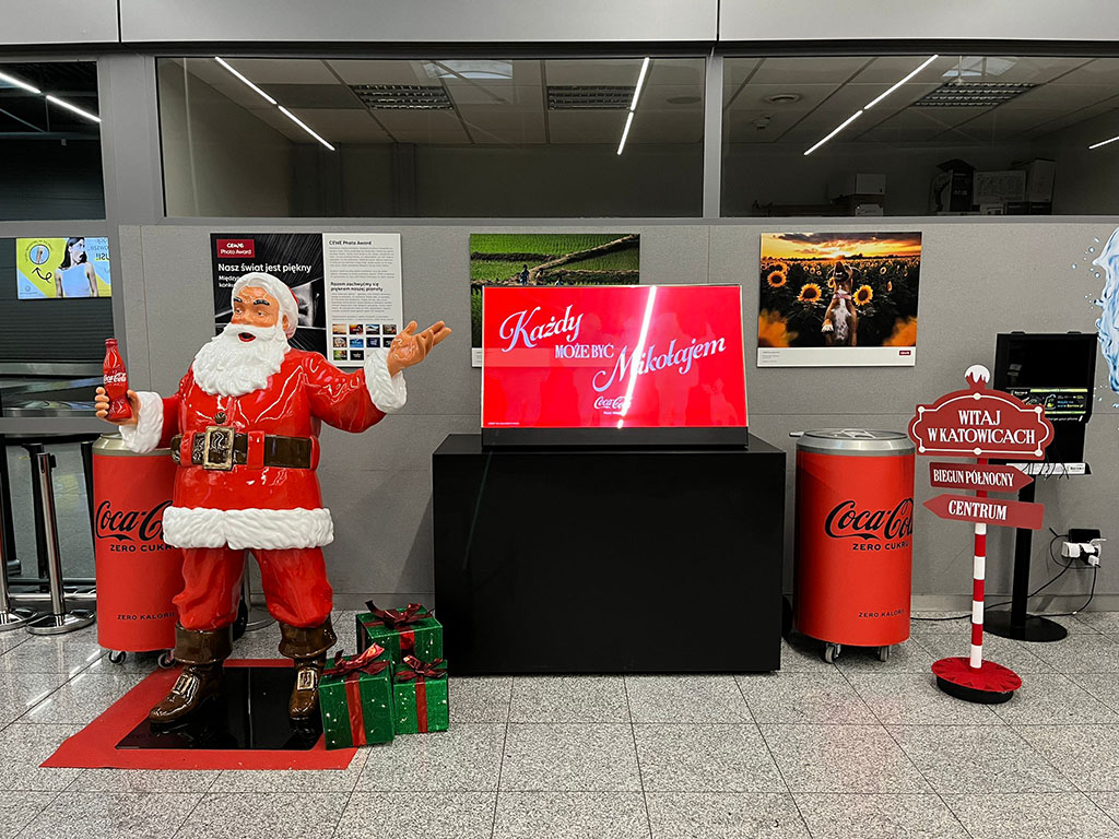 Jeder kann der Weihnachtsmann sein: Coca-Colas Weihnachtskampagne mit Weihnachtsmannfiguren 12 Figurka świętego Mikołaja w czerwonym ubraniu trzymająca butelkę Coca-Coli, stojąca obok dwóch zielonych prezentów z czerwonymi kokardami, na tle szarej ścianki z plakatami i czerwonych pojemników na napoje Coca-Cola Zero, oraz czerwony znak z napisami witającymi w Katowicach i wskazującymi kierunki.