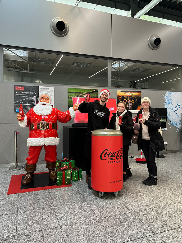 Jeder kann der Weihnachtsmann sein: Coca-Colas Weihnachtskampagne mit Weihnachtsmannfiguren 13 Trzy osoby stoją obok dużej figury Mikołaja w czerwonym stroju trzymającego butelkę Coca-Coli, każda z nich trzyma puszkę Coca-Coli, obok stoi duża puszka Coca-Coli na kółkach, tło to wnętrze budynku z szarymi ścianami i gablotami ze zdjęciami.
