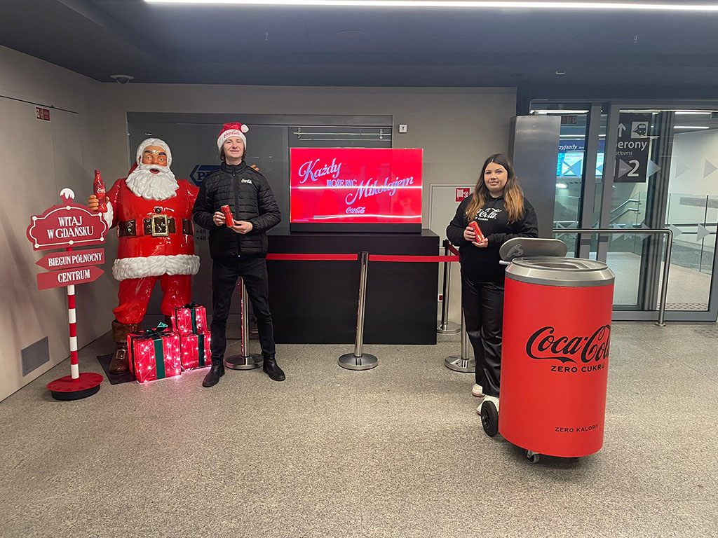 Jeder kann der Weihnachtsmann sein: Coca-Colas Weihnachtskampagne mit Weihnachtsmannfiguren 14 Dwoje ludzi trzymających puszki Coca-Coli stoi obok dużej czerwonej figurki Mikołaja i czerwonego śmietnika w kształcie puszki Coca-Coli w jasnym, nowoczesnym wnętrzu na tle ekranu z napisem w języku polskim i czerwonym stojakiem z napisem powitalnym.