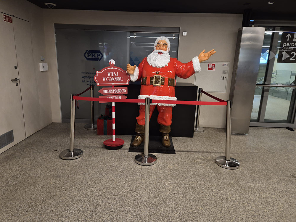 Jeder kann der Weihnachtsmann sein: Coca-Colas Weihnachtskampagne mit Weihnachtsmannfiguren 15 Figurka świętego Mikołaja w czerwonym stroju z białym futerkiem stojąca na dworcu w Gdańsku, obok czerwonego stojaka z napisem witającym w Gdańsku i kierunkami do Bieguna Północnego i Centrum, otoczona barierkami z czerwonym paskiem.