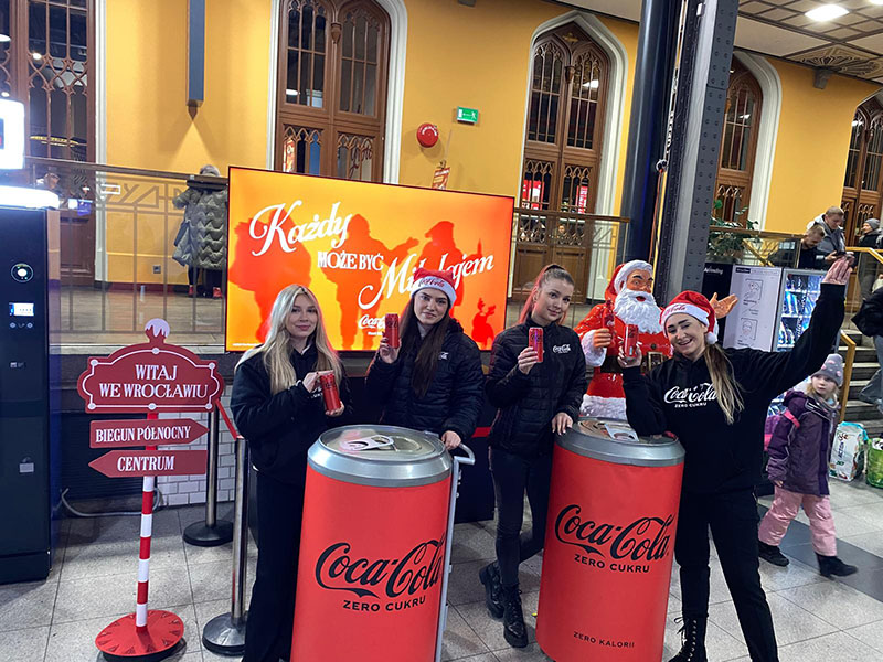 Jeder kann der Weihnachtsmann sein: Coca-Colas Weihnachtskampagne mit Weihnachtsmannfiguren 16 Cztery młode kobiety w ciemnych kurtkach i czapkach Mikołaja pozują z puszkami Coca-Coli przy dwóch dużych stojakach w kształcie puszek Coca-Coli Zero Cukru, na tle ekranu z napisem