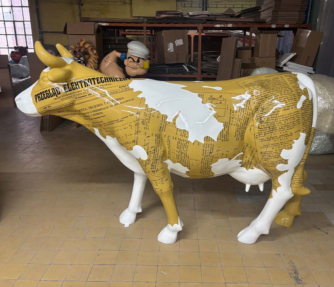 Elektrische Kuh – Symbol der Schlesischen Technischen Universität 3 Biała i żółta rzeźba krowy pokryta tekstem i schematami technicznymi, w tle figurki postaci z kreskówek oraz stosy kartonów w magazynie.