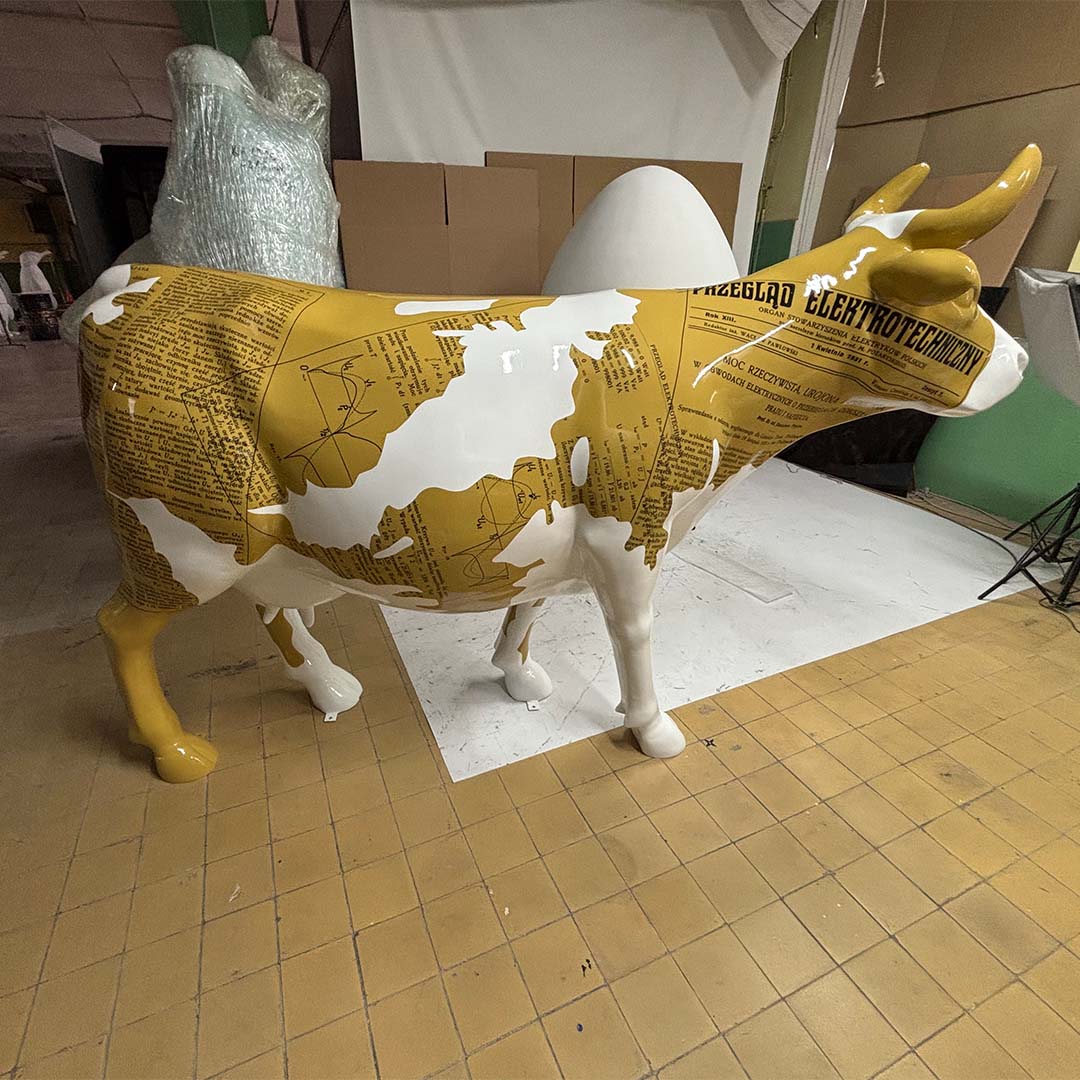 Elektrische Kuh – Symbol der Schlesischen Technischen Universität 4 Figura krowy stojąca na żółto-brązowych kafelkach, pokryta białymi i żółtymi wzorami przypominającymi strony tekstu z polskimi napisami.