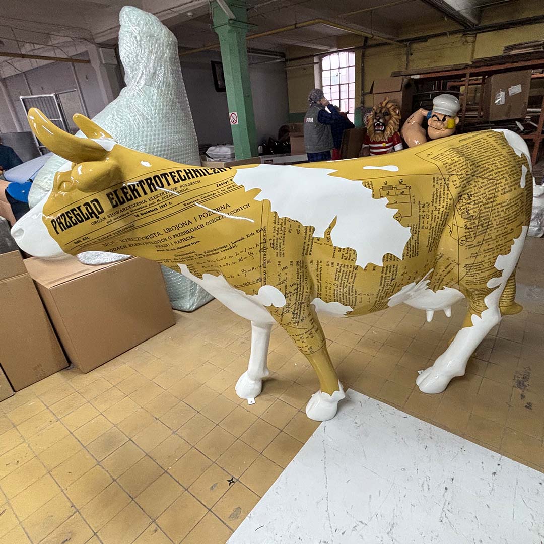 Elektrische Kuh – Symbol der Schlesischen Technischen Universität 5 rzeźba krowy ozdobiona wzorami przypominającymi stare gazety i teksty techniczne, stojąca w pomieszczeniu magazynowym z osobami i dużymi figurami w tle