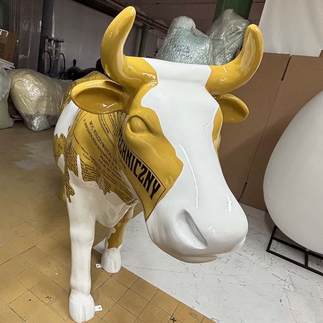 Elektrische Kuh – Symbol der Schlesischen Technischen Universität 1 Rzeźba krowy w kolorze białym i żółtym z nadrukiem tekstu, stojąca na podłodze w pomieszczeniu magazynowym lub warsztatowym, w tle kartony i owinięte folie ochronne