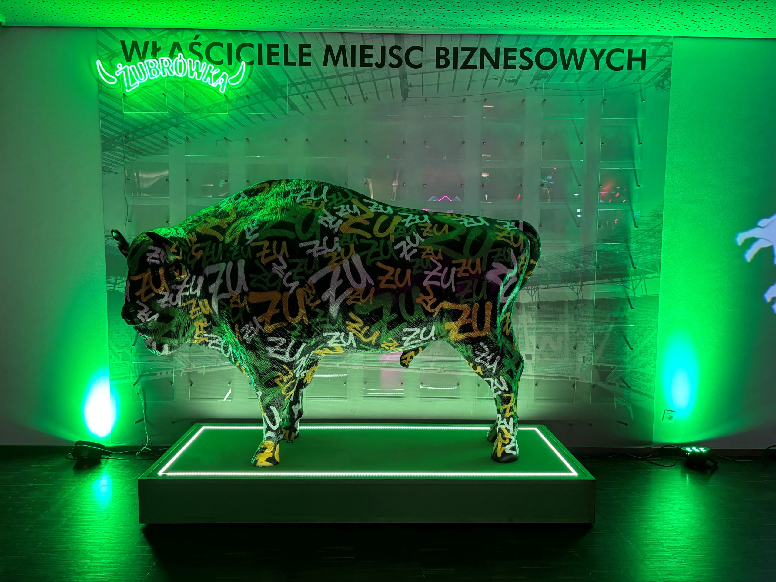 Wisent für die Wisent-Zone 3 Figurka byka pokryta graffiti z napisem 'ZU' na zielonym podświetlonym podium, z neonowym napisem Żubrówka powyżej i czarnym napisem 'WŁAŚCICIELE MIEJSC BIZNESOWYCH' na ścianie w tle, oświetlona zielonym światłem.