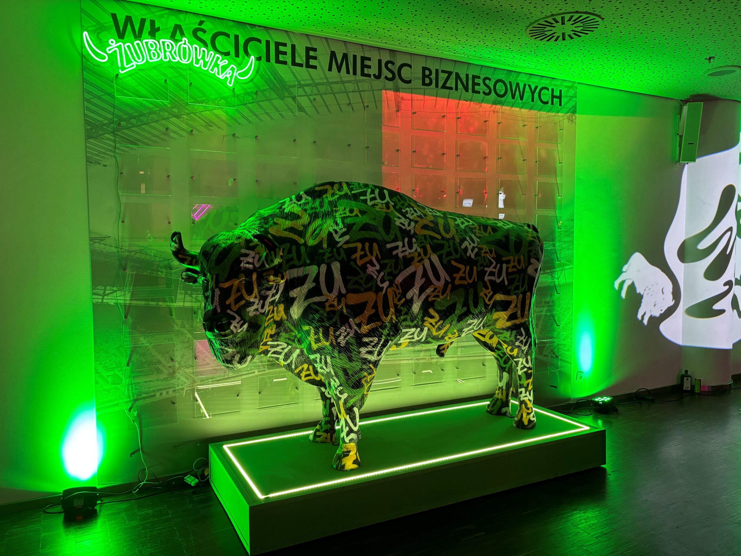 Wisent für die Wisent-Zone 1 Rzeźba bizona pokryta kolorowymi napisami „ZU” na podświetlanej zielonym światłem platformie, z neonowym napisem Żubrówka i napisem na ścianie